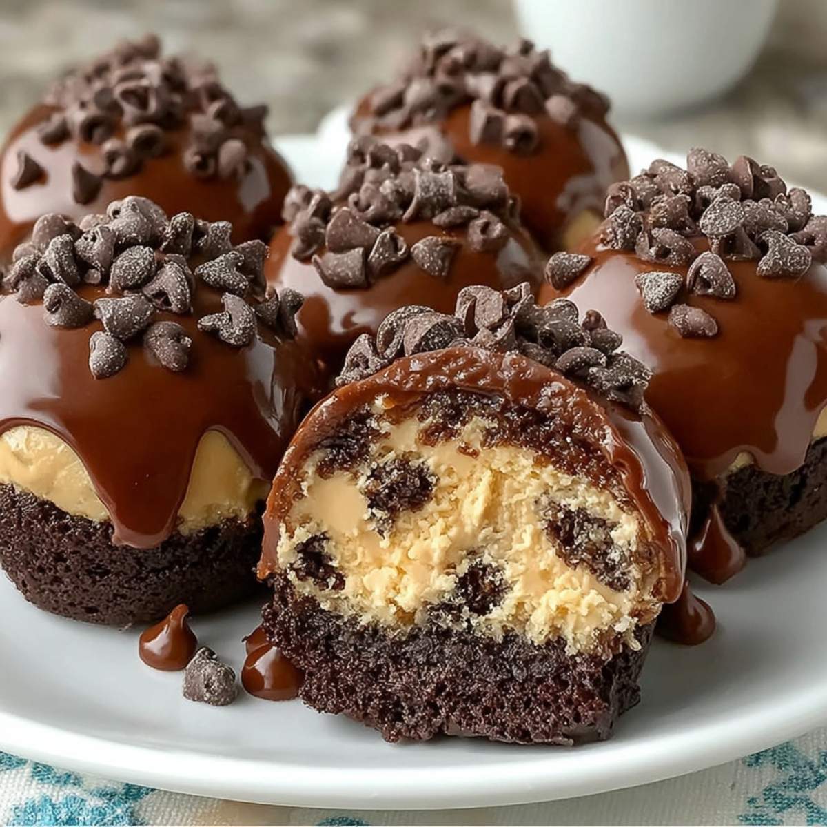 A plate of Brownie Cookie Dough Bombs, one cut open to showcase its creamy cookie dough filling, brownie layer, and glossy chocolate glaze