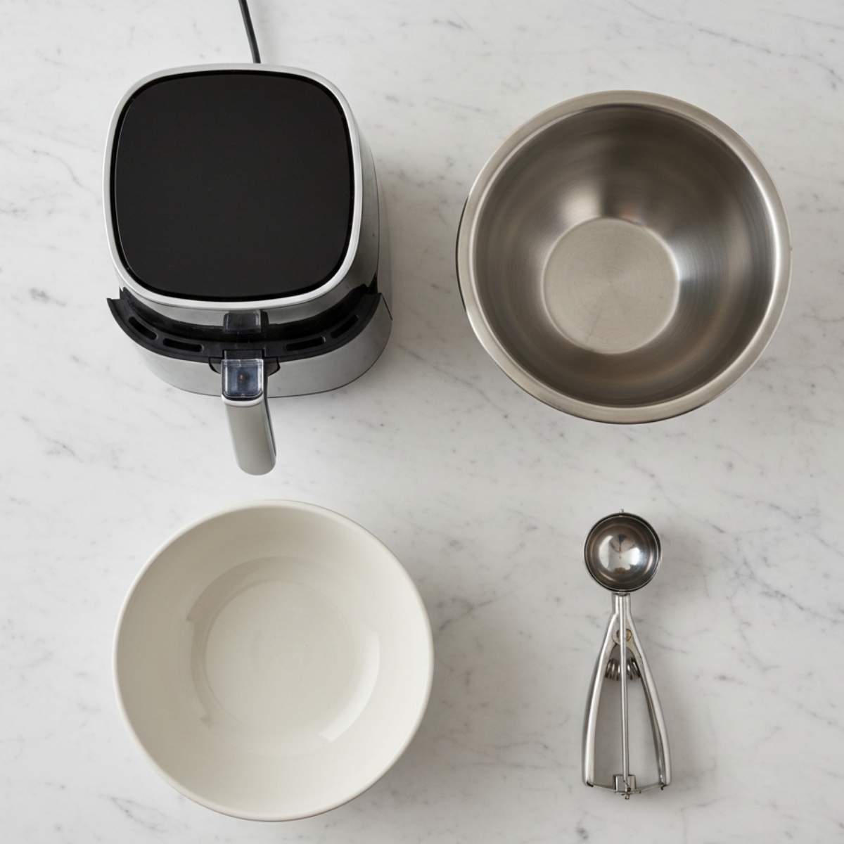 Banana donut holes equipment including an air fryer, mixing bowls, and cookie scoop arranged on a marble countertop for easy prep.