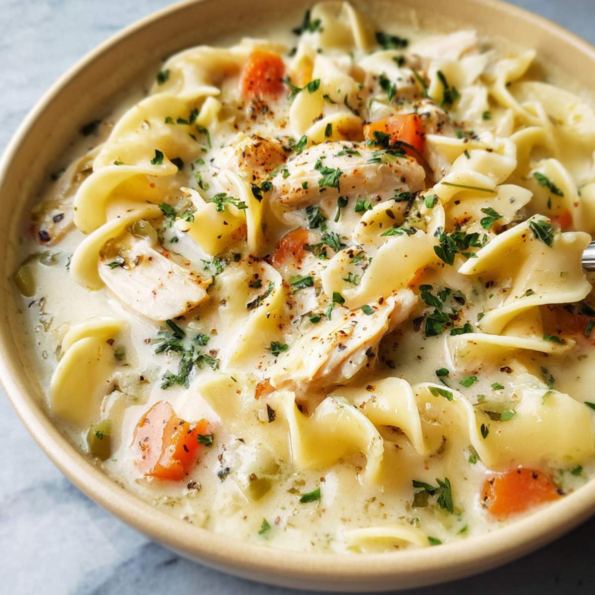 Close-up of creamy chicken noodle soup with tender egg noodles, shredded chicken, carrots, herbs, and cracked pepper in a bowl.