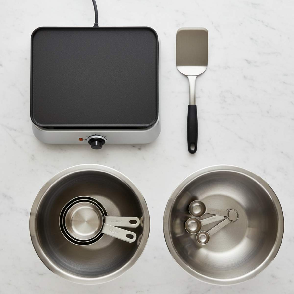 fluffy pancakes equipment including electric griddle, mixing bowls, measuring cups, measuring spoons, and spatula on marble surface.
