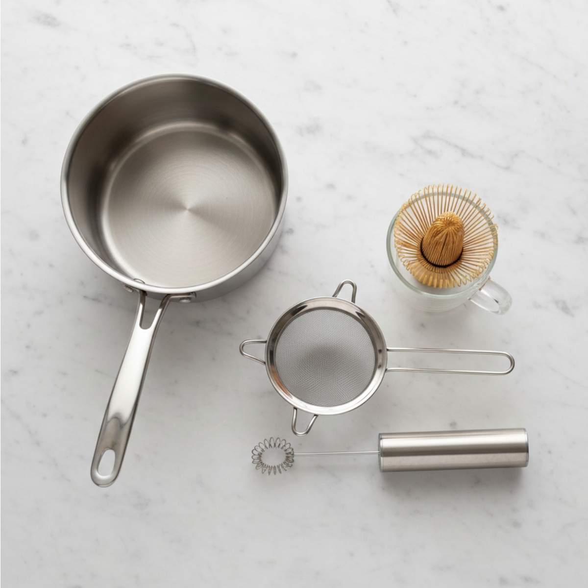 Matcha White Hot Chocolate equipment including saucepan, matcha whisk, fine mesh strainer and milk frother on marble surface.