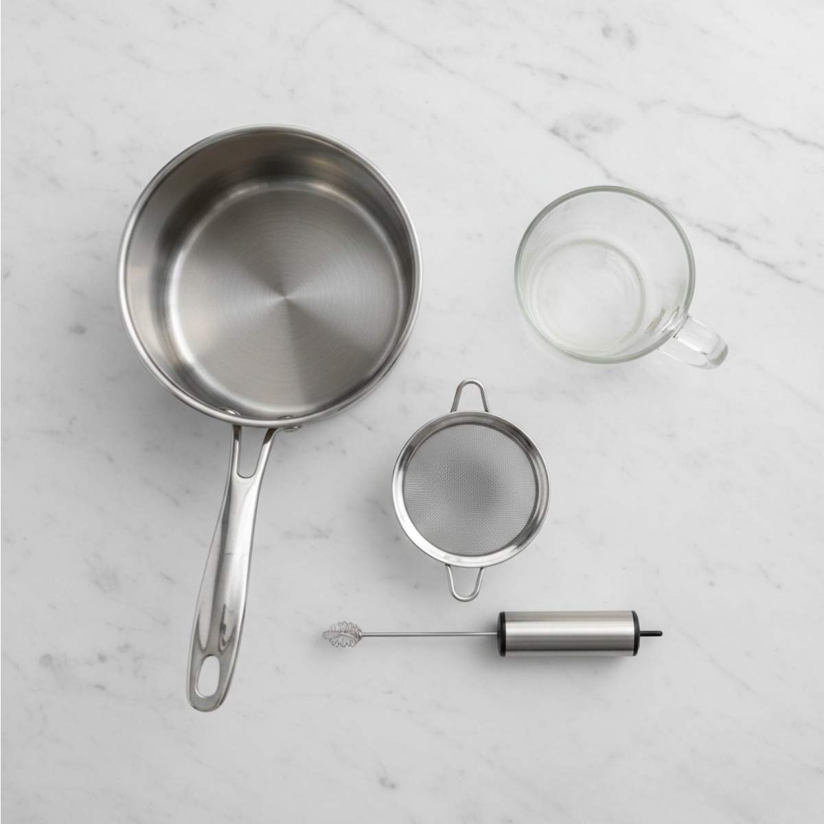 Equipment for vanilla cinnamon milk tea including saucepan, glass mixing bowl, tea mugs, and foil arranged on a marble surface.